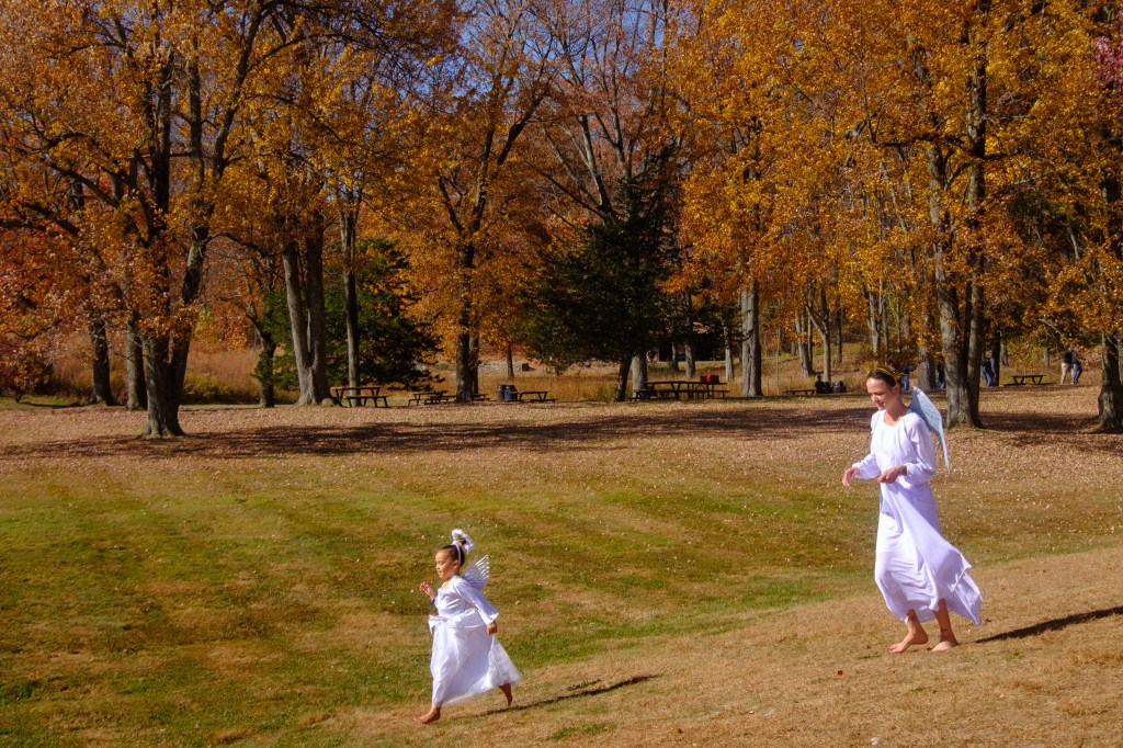 Brittany Crain and her daughter Iyla Moss dressed as angels running through the meadow area of ​​the Storm King Arts Center with fall foliage in the background